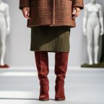 A model wearing a textured tweed coat and suede boots in an autumn runway setting with blurred mannequins and draped fabrics in warm earth tones.