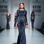 Model in a modern gown posing on a fashion week runway with stage lighting and blurred audience in the background.